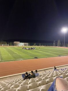 people are sitting on the bleachers at night watching something in the distance with bright lights