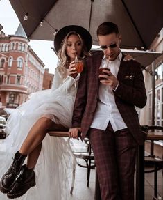 a man and woman sitting on a bench under an umbrella, one holding a drink