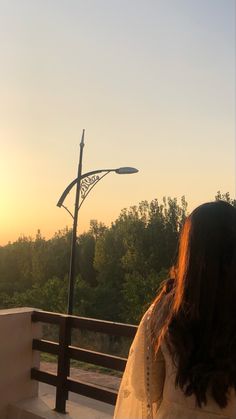 a woman standing on a balcony looking at the sun setting in the distance with trees behind her