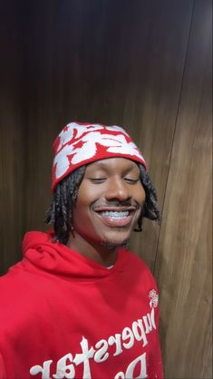 a man with dreadlocks and a red hoodie smiles at the camera while standing in front of a wooden wall