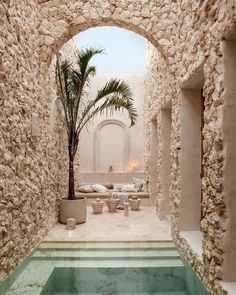 an indoor swimming pool surrounded by stone walls and palm tree in the center, with seating area on either side