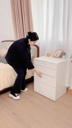 a woman in black shirt and white shoes standing next to a bed with drawers on it