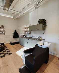 A black shampoo system with a white bowl stands in front of a large sage green cabinet unit. Boho Salon Shampoo Area, Shampoo Bowl Area Beauty Salons, Shampoo Area In Salon, Shampoo Bowl Ideas Salon, Boho Hair Salon Shampoo Area, At Home Salon Ideas, Salon Shampoo Bowl With Hair Station, Salon Lofts