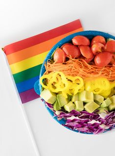 a rainbow colored plate with noodles and vegetables on it, next to a rainbow flag