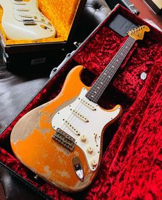 an orange electric guitar in a case on top of a red velvet covered couch next to a white and black guitar