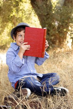 a young boy is sitting in the grass holding a red book over his face and smiling