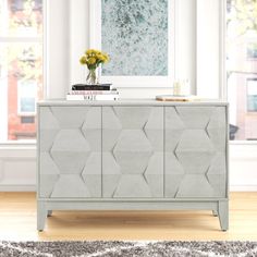 a white and grey cabinet with flowers on top in front of a large window next to a rug