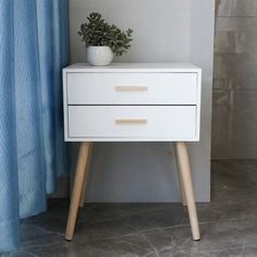 a white nightstand with two drawers and a potted plant on it next to a blue curtain