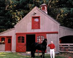 a man standing next to a black horse in front of a barn with red doors