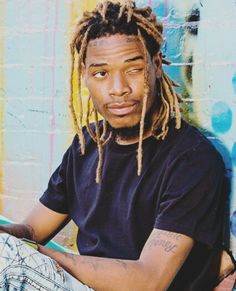 a man with dreadlocks sitting in front of a graffiti wall holding a skateboard