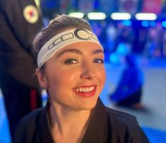 a woman with a blindfold on her head smiles at the camera while another person stands in the background