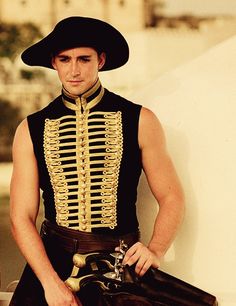 a man wearing a black hat and uniform with gold trimmings, standing next to a wall