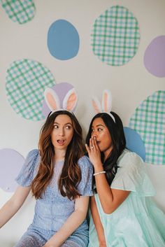 two young women sitting next to each other in front of a wall with bunny ears on it