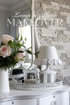 a white dresser topped with a vase filled with flowers next to a mirror and lamp