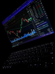 an open laptop computer sitting on top of a black table with a monitor displaying stock market data