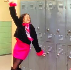 a woman in a pink skirt and black jacket standing next to lockers with her arms up