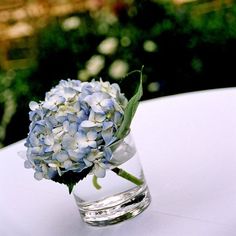 a small glass vase filled with blue flowers on top of a white table cloth covered table