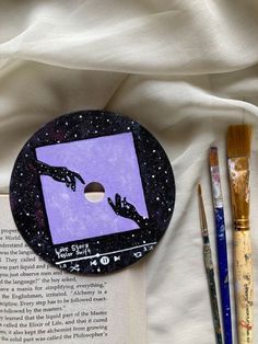 an open book next to some paint and brushes on a white sheet with a purple background