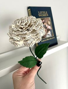 a person is holding a flower made out of old book pages and green leaves in front of a bookshelf