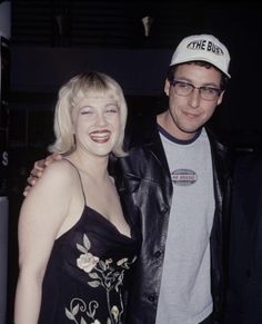 a man standing next to a woman in a black dress and white baseball cap at an event