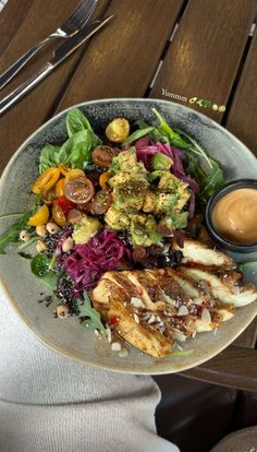 a person holding a plate with chicken, salad and dressing on it in front of a wooden table