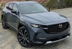 the front end of a gray suv parked on gravel with mountains in the back ground