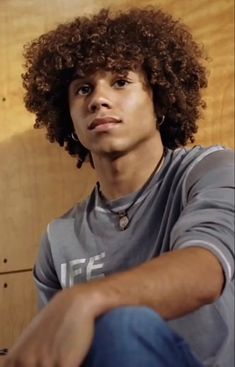 a young man with curly hair sitting in front of a wooden wall