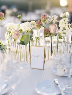 the table is set with white and pink flowers in vases, place cards and silverware