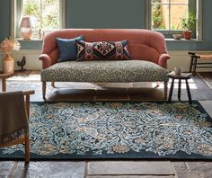 a living room filled with furniture and pillows on top of a blue rug in front of two windows