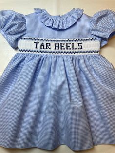 "This cute hand smocked little girl's dress is made of pale blue 1/16\" 100% cotton gingham. Tar Heels, your school's mascot name, or the school's name are smocked to match the fabric on the white insert, (maximum 9 letters). A ruffled neckline and short puff sleeves finish the dress. On the back are three buttons and a belt. It has a generous 3-4 inch hem. This can also be made in other colors or fabrics. Also, a little boy's shortall or bubble to match can be made. If you'd like a different co Southern Baby Clothes, Football Gameday Outfit, North Carolina Football, Carolina Football, Southern Baby, Hand Smock, Ruffled Neckline, Gingham Fabric, Tar Heels