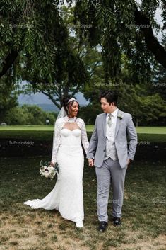 a bride and groom holding hands walking through the grass in front of some large trees