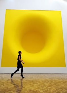 a woman is walking in front of a large yellow art piece on the wall behind her