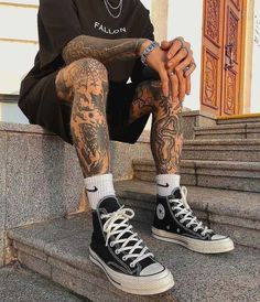 a man with tattoos sitting on the steps wearing black converses and holding his hands in his pockets