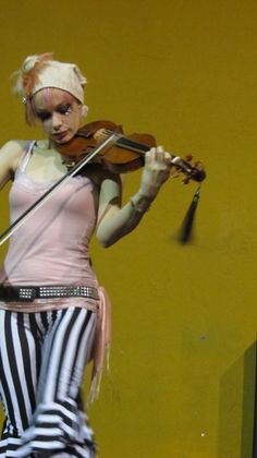 a woman with white hair playing violin on stage in front of a yellow wall and black and white striped pants