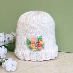 a white knitted hat with flowers and leaves on the side next to some flowers