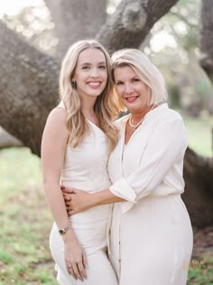 two women standing next to each other in front of a tree with their arms around each other