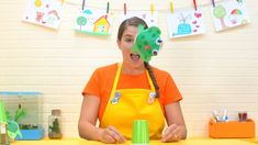 a woman wearing an orange shirt and yellow apron holding a green cup in front of her face