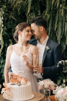 a bride and groom holding champagne glasses in front of a cake with greenery behind them