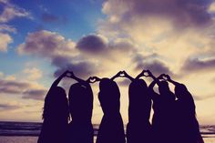 four women standing in front of the ocean making a heart shape with their hands at sunset