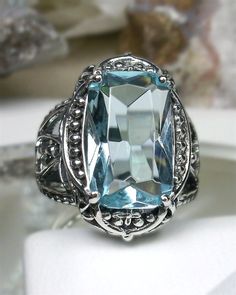 Simulated Blue Aquamarine Ring Cush Rectangle Gothic Design#142 Custom Made Lovers of Gothic and Art Deco styles will adore this stunning aquamarine blue ring. Handcrafted in solid sterling silver, this ring features a striking filigree design that adds a touch of vintage charm. The centerpiece of this ring is a brilliant 6ct simulated blue aquamarine gem, carefully placed in a rectangle-cut setting for maximum sparkle. The flawless gem measures 15mm by 9mm and is securely held in place by the i Blue Topaz Ring With Square Cut, Blue Topaz Ring With Square Cut Gemstone, Blue Square Cut Topaz Ring Gift, Gift Blue Square Cut Topaz Ring, Elegant Rectangular Blue Sapphire Ring, Blue Aquamarine Jewelry With Rectangular Stone, Blue Topaz Square Cut Jewelry, Blue Rectangular Topaz Ring With Accent Stones, Rectangular Blue Topaz Ring With Accent Stones