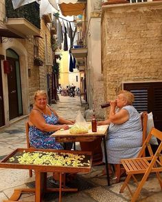 two women sitting at a table with food in front of them on an alleyway