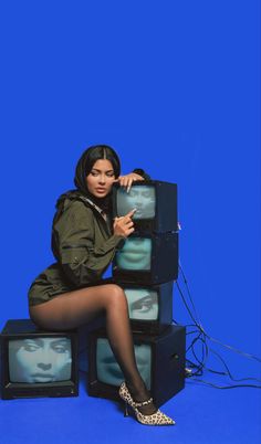 a woman sitting on top of four televisions in front of a blue background with her legs crossed