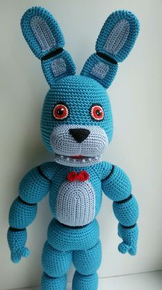 a crocheted blue stuffed animal wearing a red bow tie and standing on a white surface