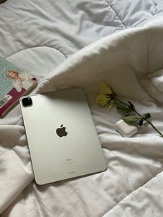 an apple ipad on a bed next to a book and a vase with flowers in it