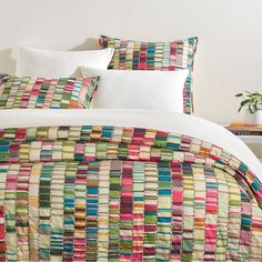 a bed with white pillows and colorful quilts on it's coverlet, next to a small potted plant