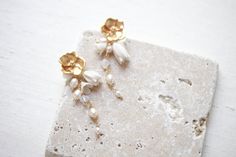 two gold and white flower earrings sitting on top of a stone slab with pearls hanging from it