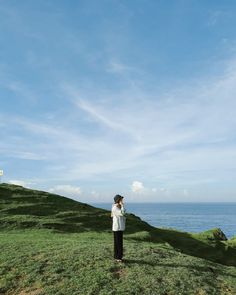 a man standing on top of a lush green hillside next to the ocean with a cell phone in his hand