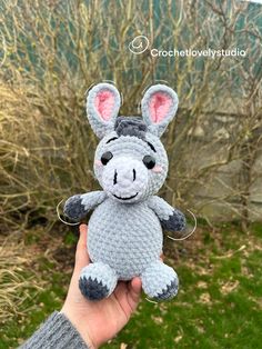 a hand holding a small gray and white crocheted stuffed animal in front of some bushes