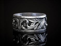 an intricately designed silver ring sits on a reflective surface in front of a black background