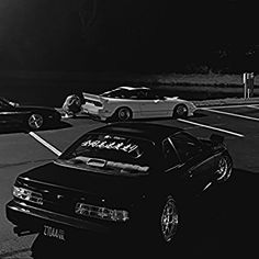 black and white photograph of two cars parked in a parking lot at night with people standing on the sidewalk